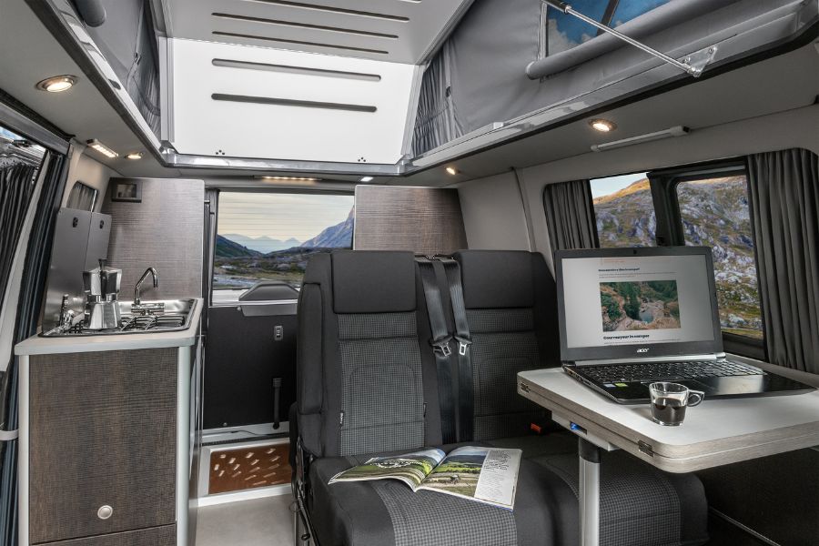 Postazione di lavoro allestita nella dinette di un furgone camperizzato, con laptop, tazza di caffè e vista sulle montagne dal finestrino