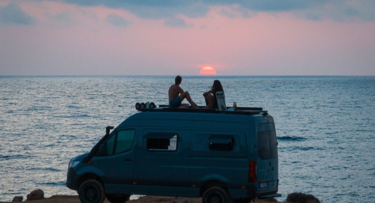 Furgone camperizzato al tramonto in riva al mare, coppia seduta sul tetto: lo stile di vita di chi fa smart working in camper